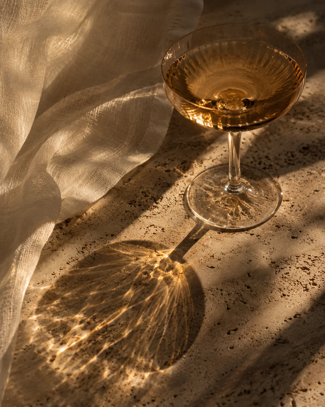 Amber coupe glass casting warm reflections on travertine beside ivory gauze.