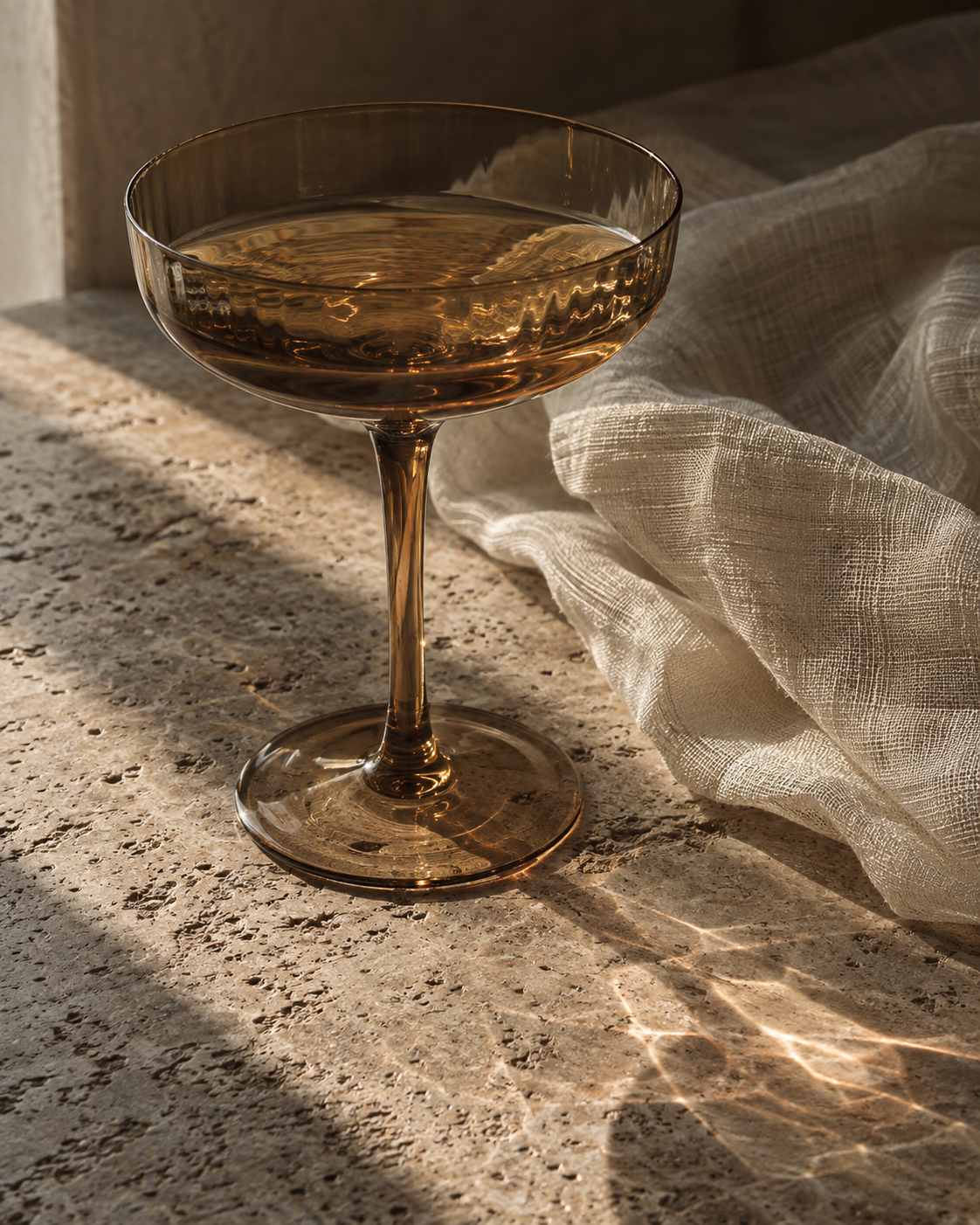 Amber coupe glass in warm sunlight on travertine beside ivory linen.