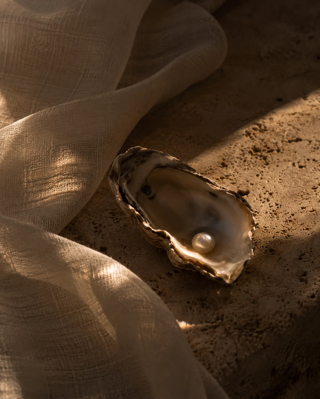 A pearl resting in a shell beside ivory linen on warm stone.